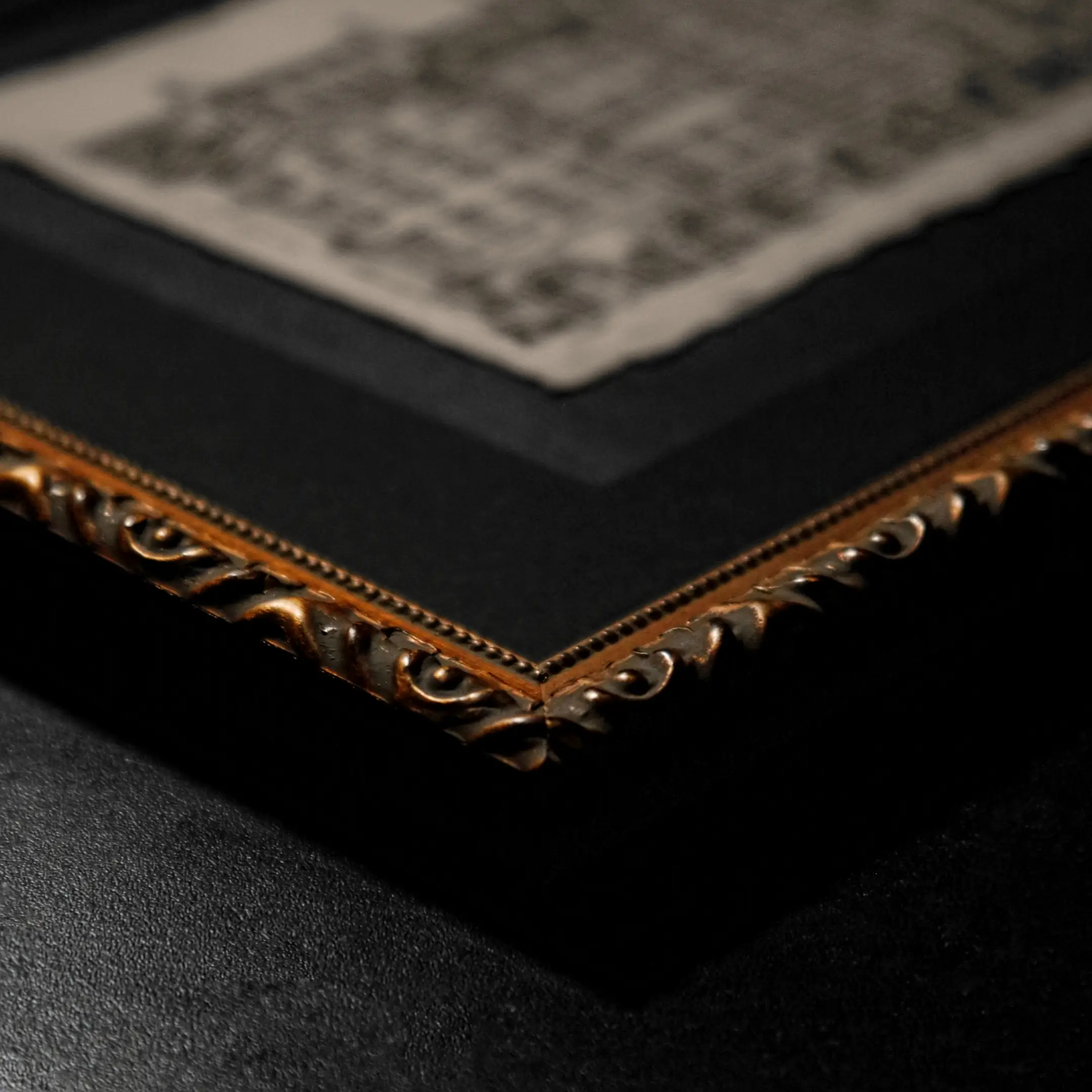 A close-up shot of an ornate, gold-toned picture frame corner with intricate carvings. In the blurred background, the ink drawing "Civitas" on aged paper is visible inside a black mat.