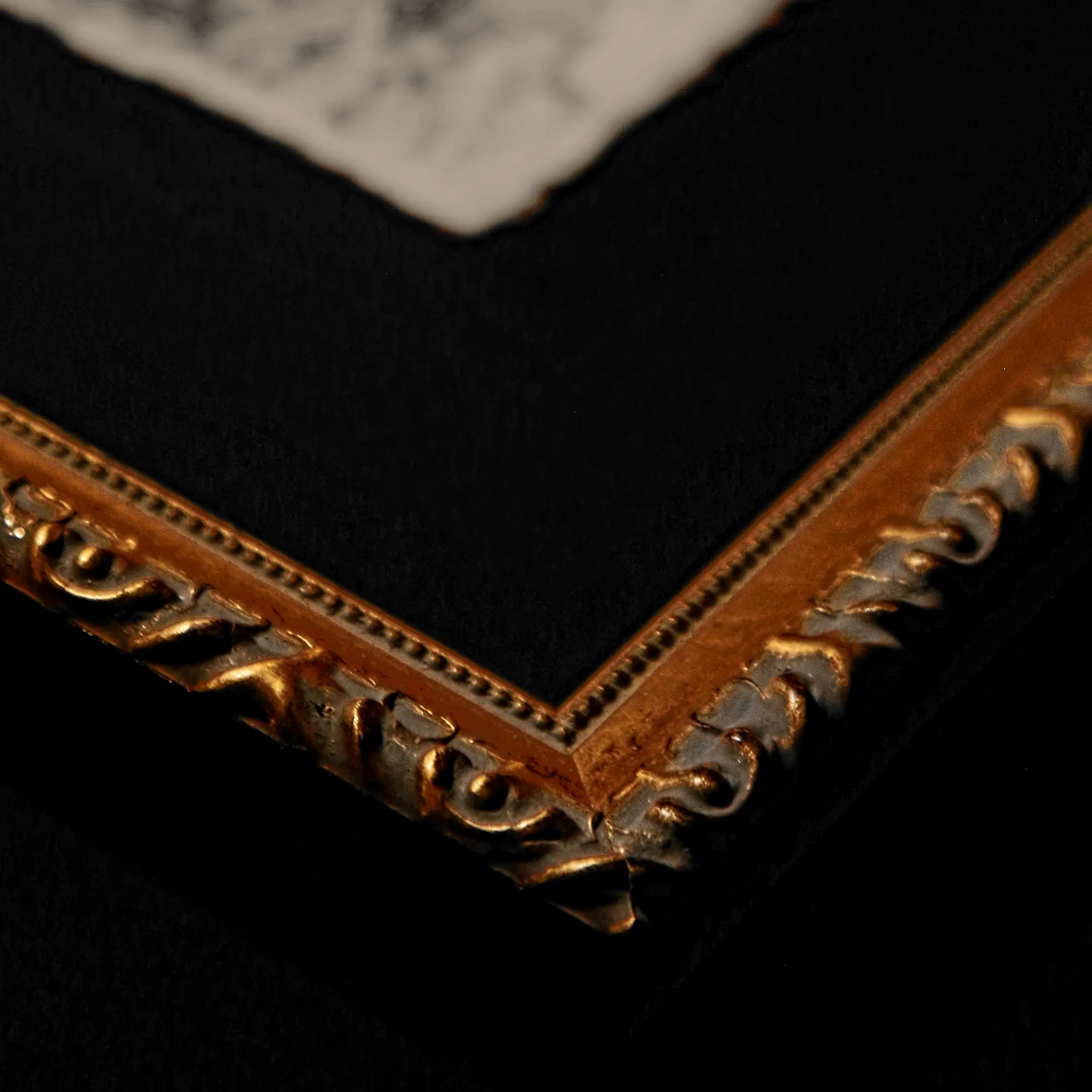 A macro close-up of the bottom corner of a gold ornate picture frame. The focus is on the intricate carved patterns of the gilded wood, the deep black velvet-like texture of the matting, and a glimpse of the deckled edge of the paper above.